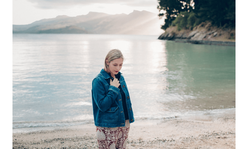 Photograph of the author Eleanor Catton, standing on a beach with a still bay behind her. She is wearing a denim jacket and looks contemplative.
