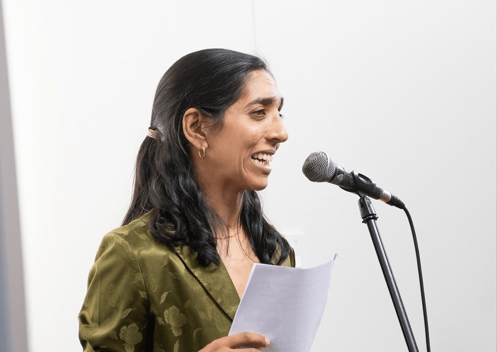 Neelu Jennings, a brown skinned woman with shoulder length dark hair, reads from a sheet of paper held in her hands while speaking into a microphone