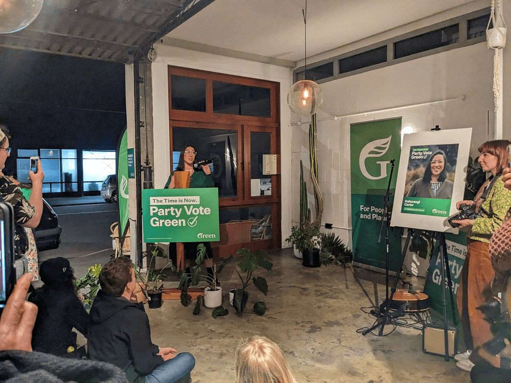 a room with a tall Māori woman wearing green and lots of party vote green signs, onlookers smiling as she speaks