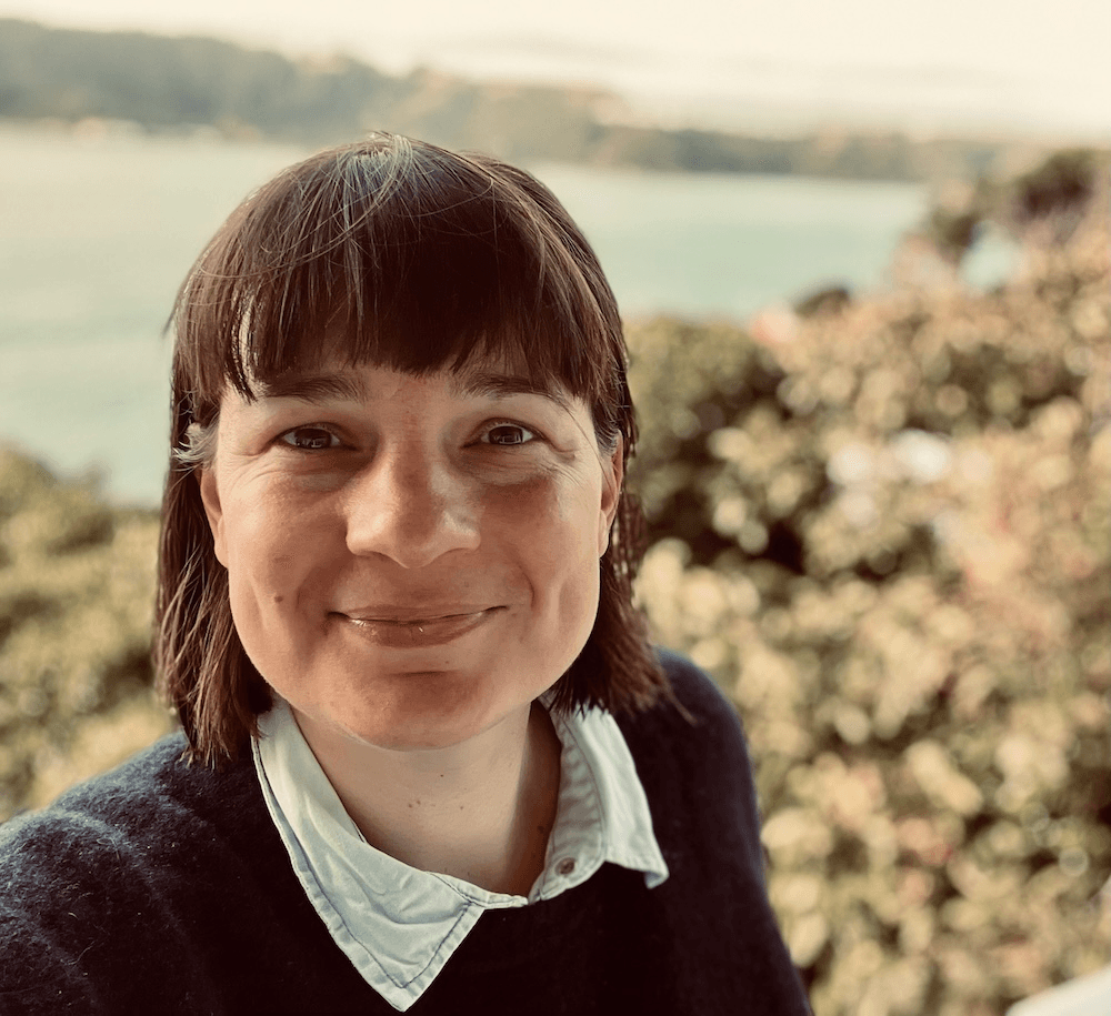 A white woman with short darck heair and collared shirt smiling at the camara with water in the background