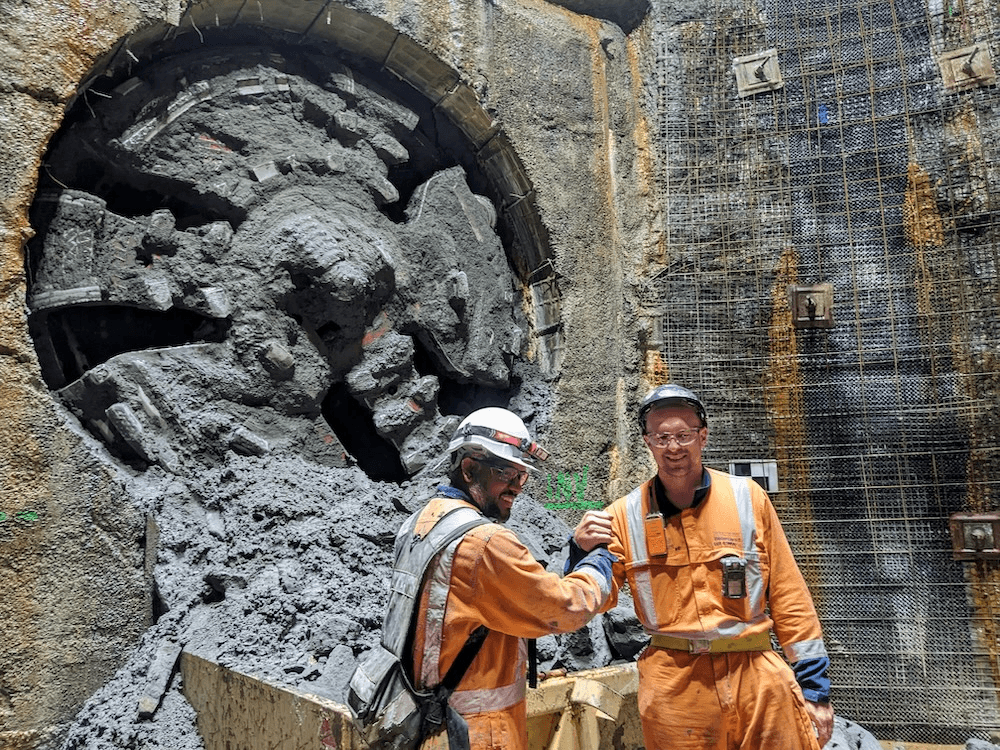 two men in orange shake hands in front of a slightly looming circular machine with lots of teeth for gnawing through rock