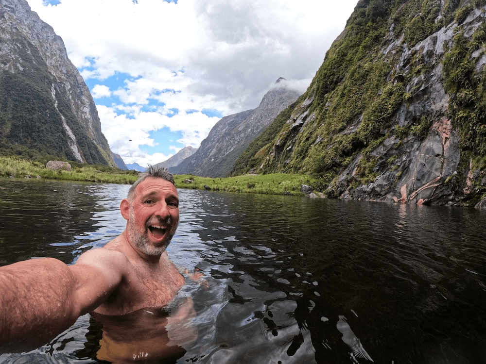 a white man with a huge beam smiles for the camera in a silky dark lake surrounded by impressive mountains
