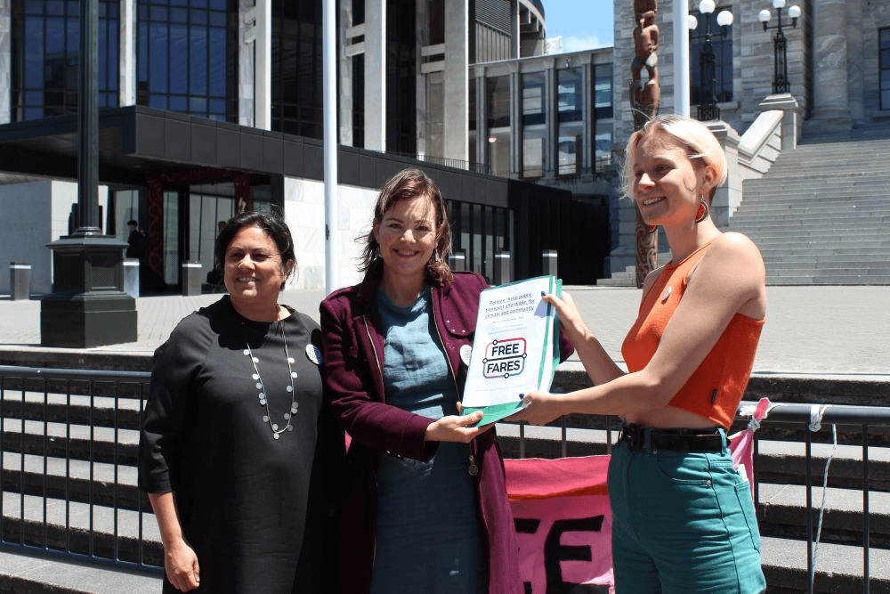 a woman with an orange shirt and platinum blonde hair smilling on the parliament forecourt holding a bit of paper with julie ann genter in a lovely maroon jacket and ayesha verral a brownskinned woman in a black dress looking in a slightly different direction
