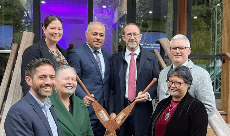Seven people, some smiling, pose in front of a building entrance. Two women in front hold carved wooden paddles and display traditional moko kauae. The group appears to be attending a formal or cultural event.