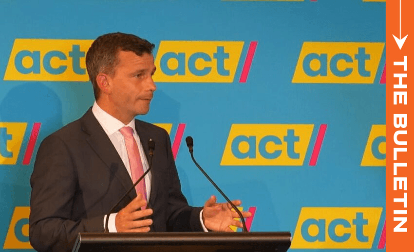 Man in a suit stands at a podium speaking into microphones. The background displays "act" repeated multiple times. An orange vertical banner on the right reads "THE BULLETIN.