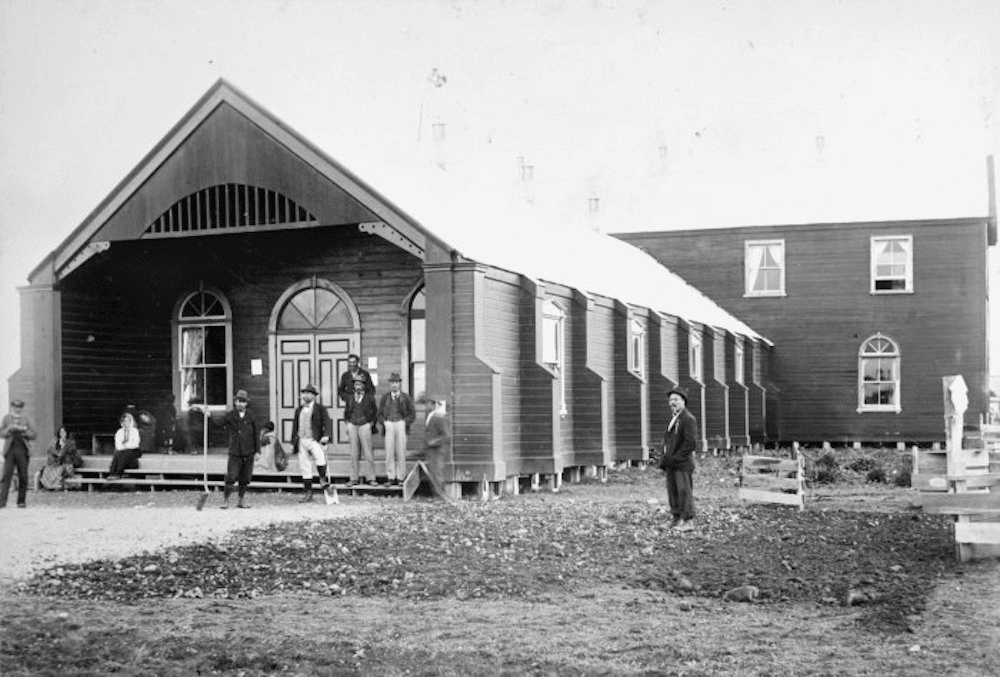 This photo shows the purpose-built wharenui in Pāpāwai hosted the sixth and seventh Te Kotahitanga hui in 1897 and 1898. The wharenui looks more similar to a Western church than the kind of wharenui we have come to expect nowadays, as it lacks carving. There are two buildings, one single story building at the forefront and another two story building behind.