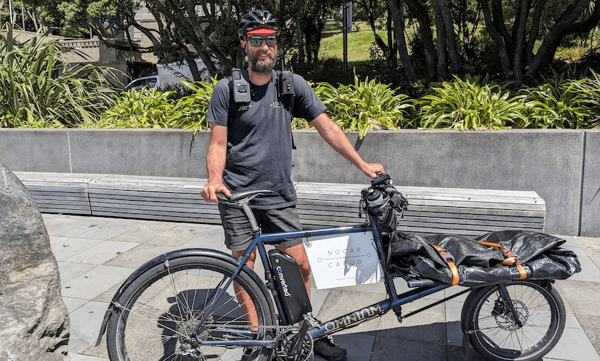 Chris baker, a smiling man with a dark grey tshirt and a helmet holding a long cargo bike