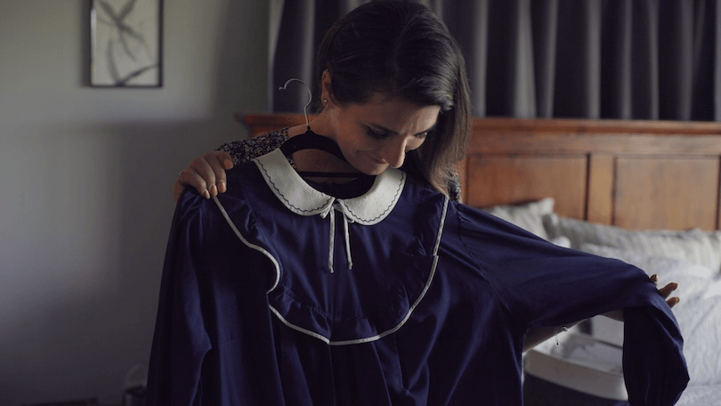 a woman in plain clothes holds the iconic gloriavale dress with a white collar and a white line of piping around the circular yoke, looking wary