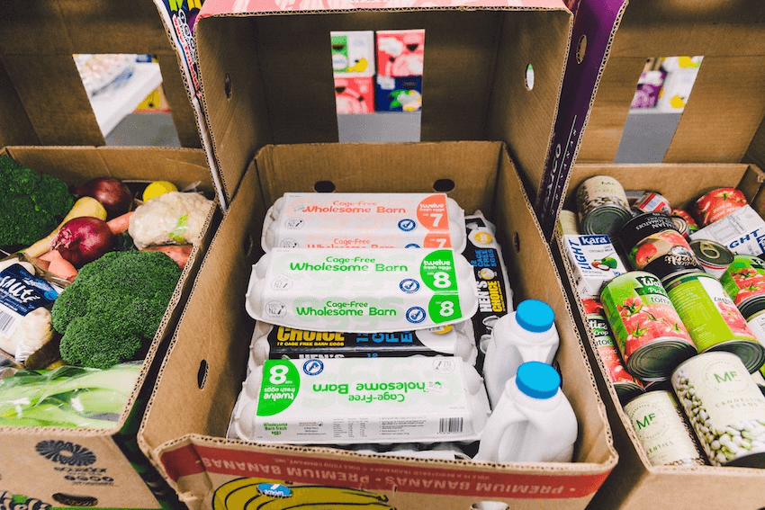 a cardboard box with eggs and milk bottle in it, light blue top. next to it are boxes with veges and canned food