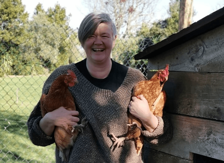a grey haired soman with short hair, a big smile and broad shoulders smiles while wearing a brown jumper and holding two golden brown chickens, with sunlit trees in the background