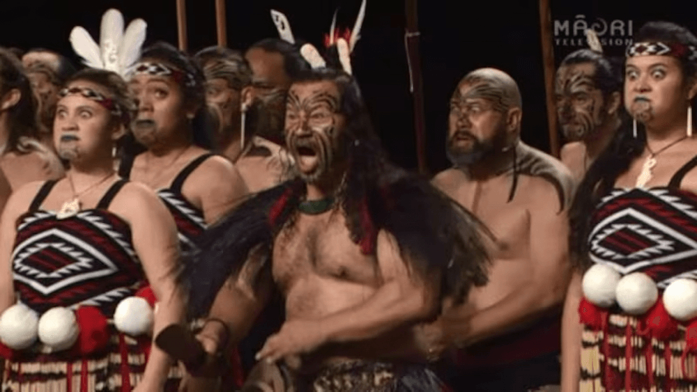 Paora Sharples performs at the 2016 Tāmaki Makaurau regional kapa haka competition alongside Te Rōpū Manutaki. They are all dressed in traditional Māori dress, some female wield poi and all performers have moko (either real tattoos are drawn on for the performance).