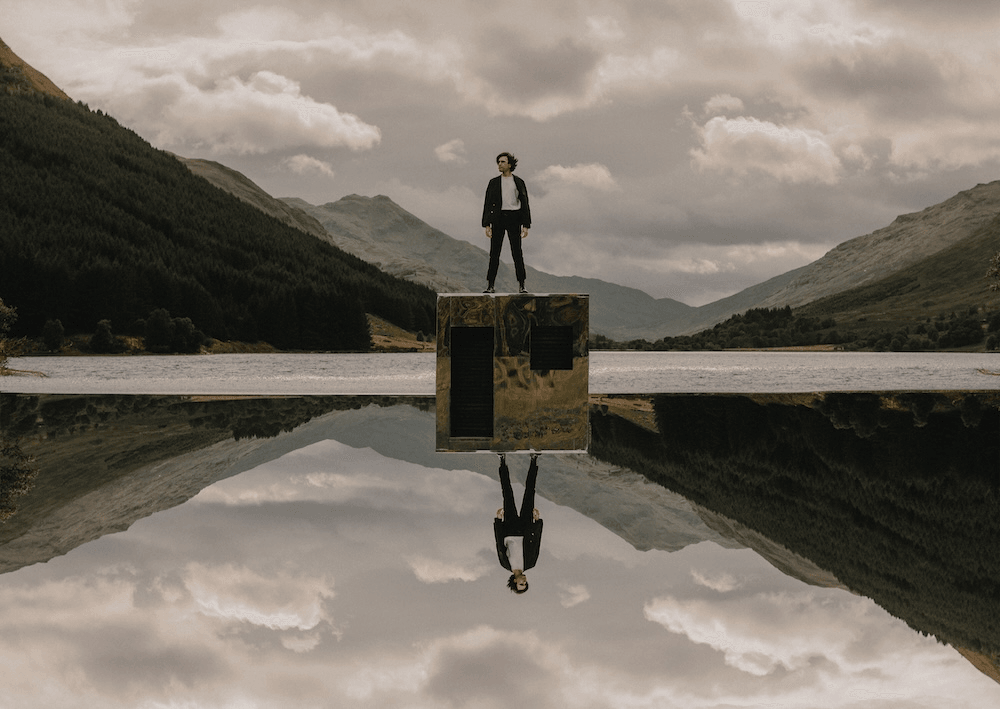 a guy in a white shirt and dark coat and jeans sanding on a shiny silver cube in a theortically scottish misty valley with his reflection beneath him