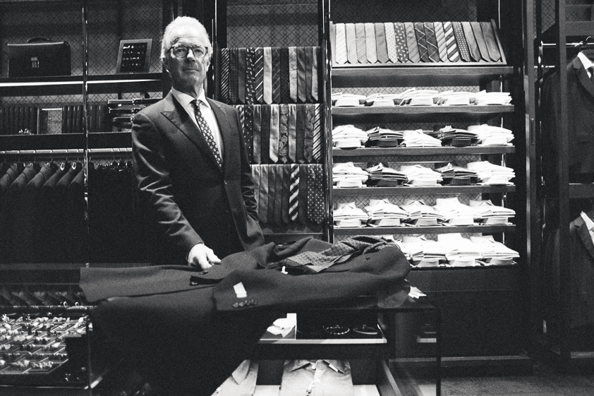 A black and white film photo of an older man wearing a suit and standing in his suit shop