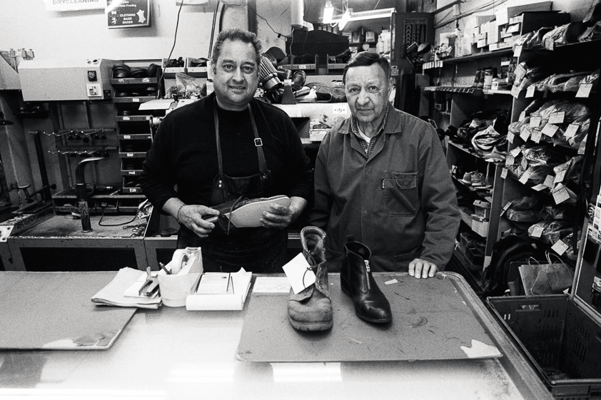 Black and white film photo of two men standing behind the counter of their shoe repair shop