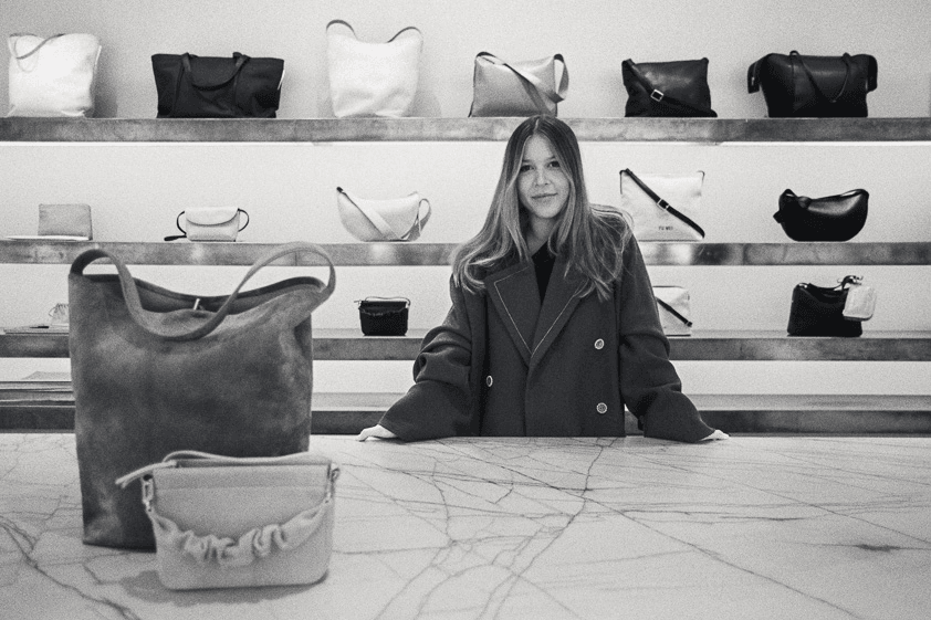 Black and white photo of a woman standing at the counter of her leather bag store with bags around her