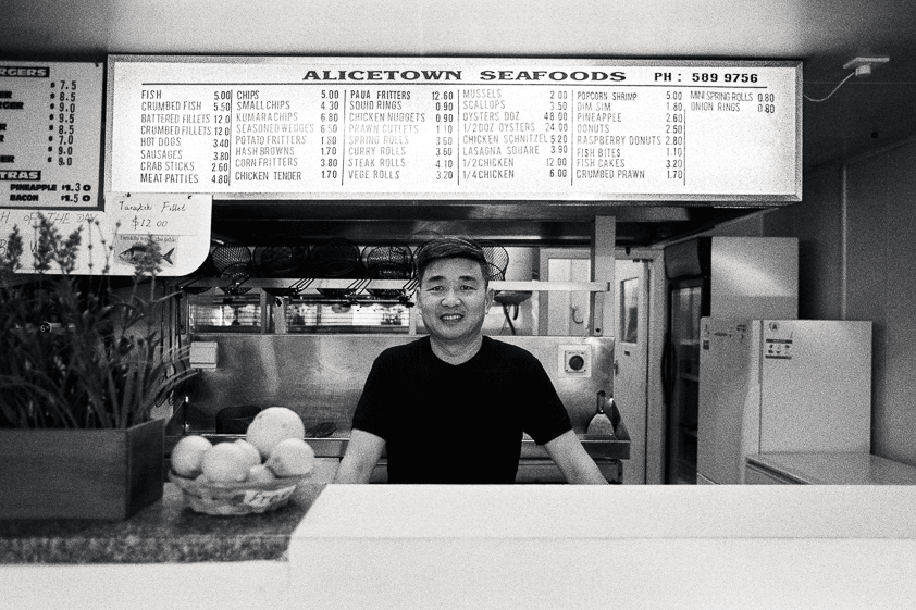صورة بالأبيض والأسود لرجل يقف خلف المنضدة في متجر السمك والبطاطا المقلية الخاص به مع قائمة فوقه وخلفه
