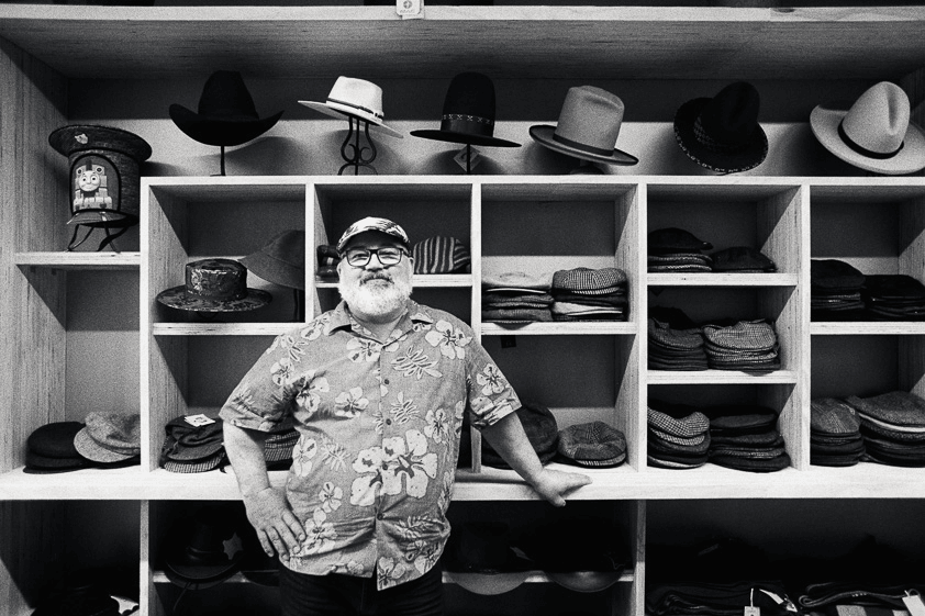 Mężczyzna z imponującą brodą w swoim sklepie z kapeluszami, kapelusze wisiały na ścianie za nim