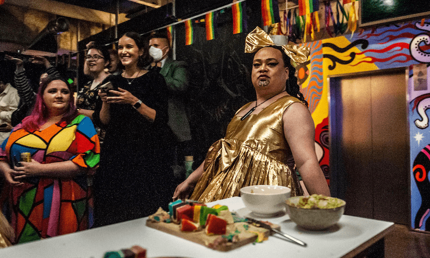 A person in a sparkly gold dress leans on a table that has birthday snacks on it and pride flags in the background. 