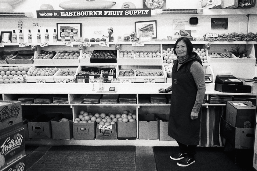 A woman stands in her fruit and vege shop, with goods behind her
