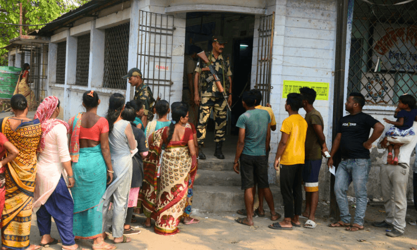 women in colourful saris line up outside a building which has soldiers in it in india, ready to vote