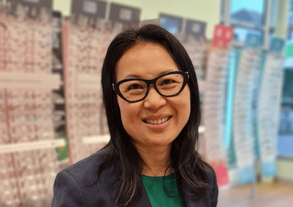May Young, an Asian woman in a specsavers store, wearing green an blue and smiling at the camera