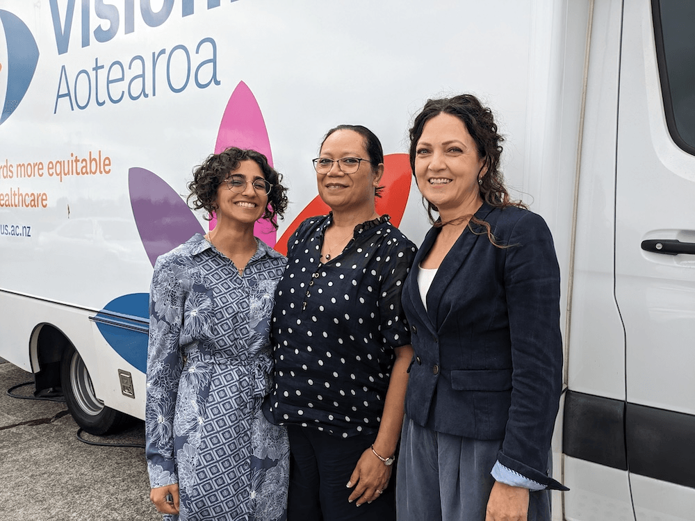 three women wearing blue tones outside a coloursful bus. Sachi, on the levt is short, with light brown skin, tightly curling hair and a big smile; Sila, in the middle is a pacific woman with straight hair tied back and rectangular glasses. Germaine, on the right, is a lebanese woman with no glasses and a friendly smile