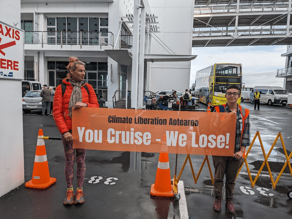 two protesters in a parking lot holding an orage sign saying 'you cruise we lose' one is tall and wearing a BRIGHT orange jacket, the other is short and wearing brown docs, a checked flannel shirt and brown boots