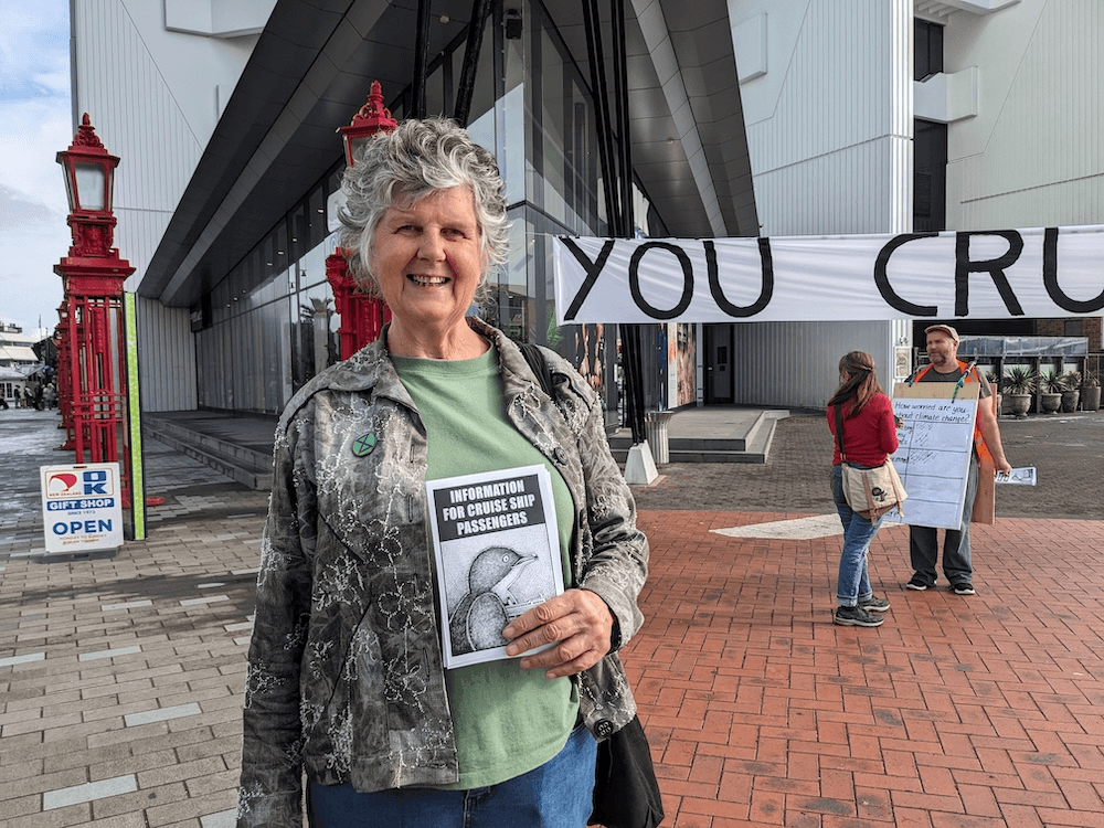 a grey haired woman with a green t-shirt and a smile holding a pamphlet with a picture of a cruise ship and a penguine 