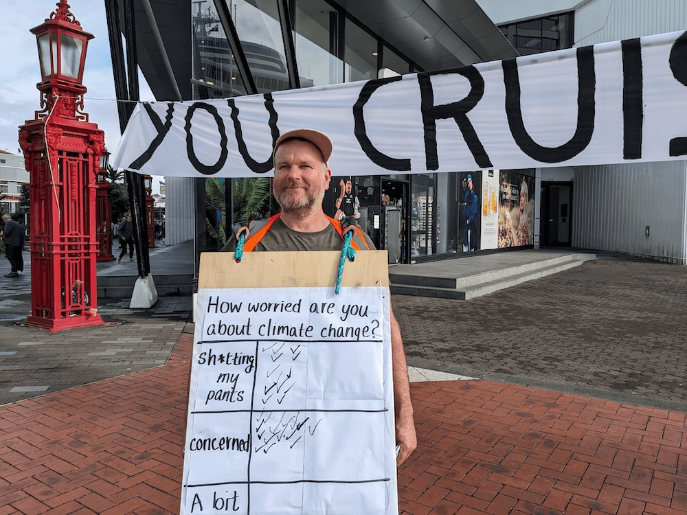 A middle aged man smiling in front of a banner wearing a sandwhich board with check marks. lots of people have checked that they are "sh*tting their pants about the climate crisis)