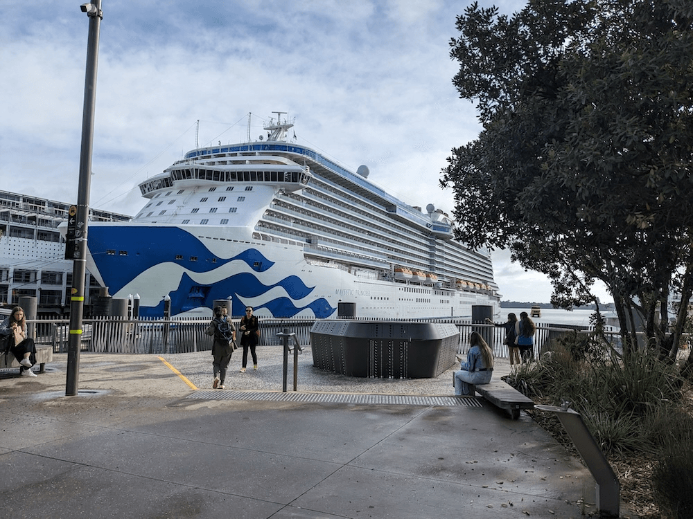 A cruise ship with blue waves on the bow is docked on the Auckland waterfront, the sky wis blue with streaks of cloud and there are people in the foreground