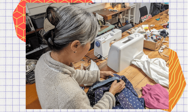 a grey haired asian woman bends over a backpack she is stitching on a table with sewing machines on it