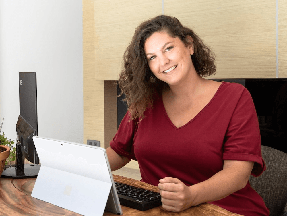 a light skinned woman with medium brown curly hair sitting in front of a folding tablet and smiling