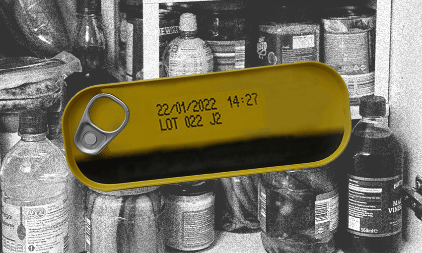 An open can of sardines or similar with a best before date stamped on the top, against a background of food items in a pantry