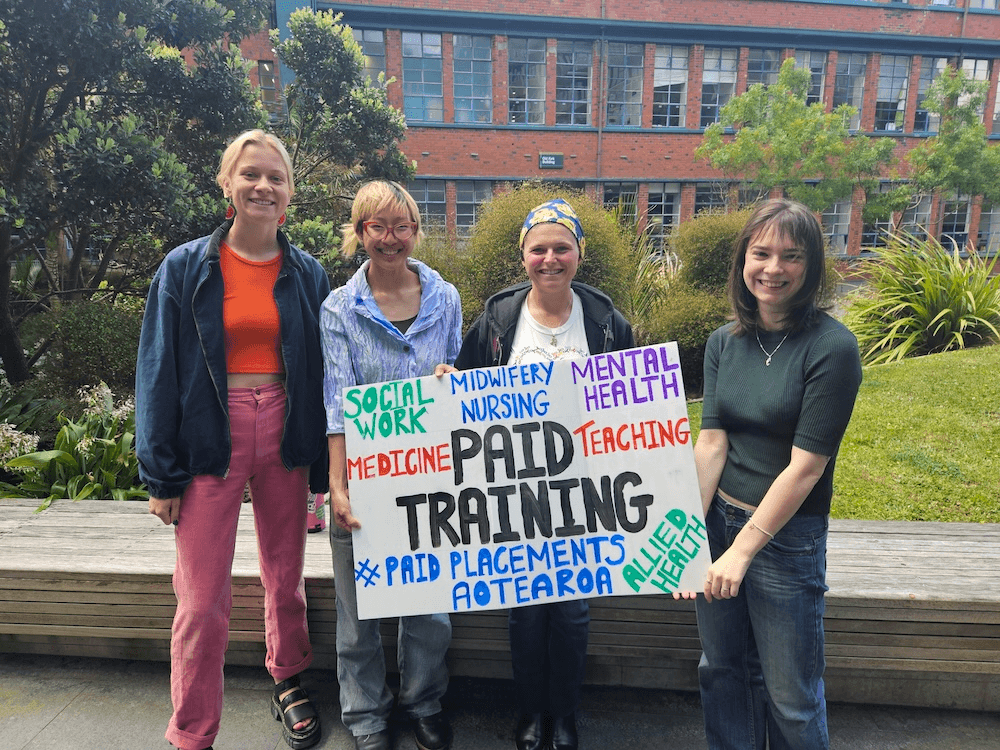 four people holding a sign saying PAID TRAINING with grass and a building in the background