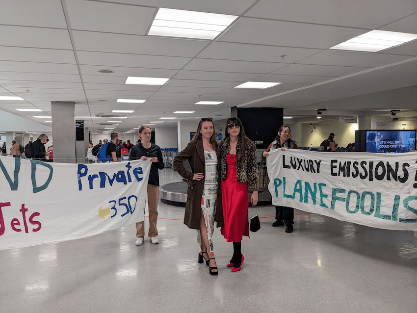 a airport baggage terminal with two women wearing silky dresses posing in front of people holding signs that say 'no private jets' and 'luxry emissions + plane foolish"
