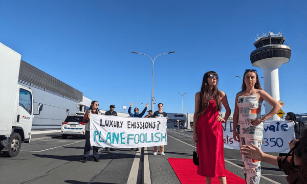 a blue sky and a red carpet with two women wearing slip dresses posing in front of an air traffic control tower with protesters holding anti private jet signs in the distance