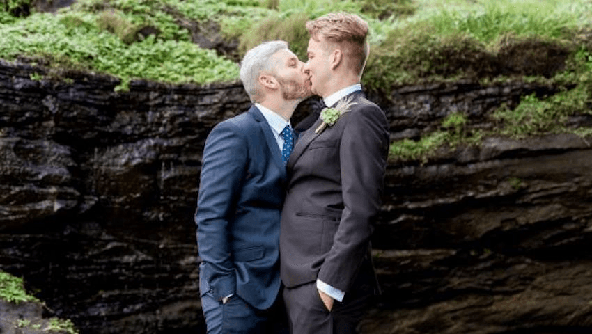 Two blonde men kiss on their wedding day against a scenic green backdrop
