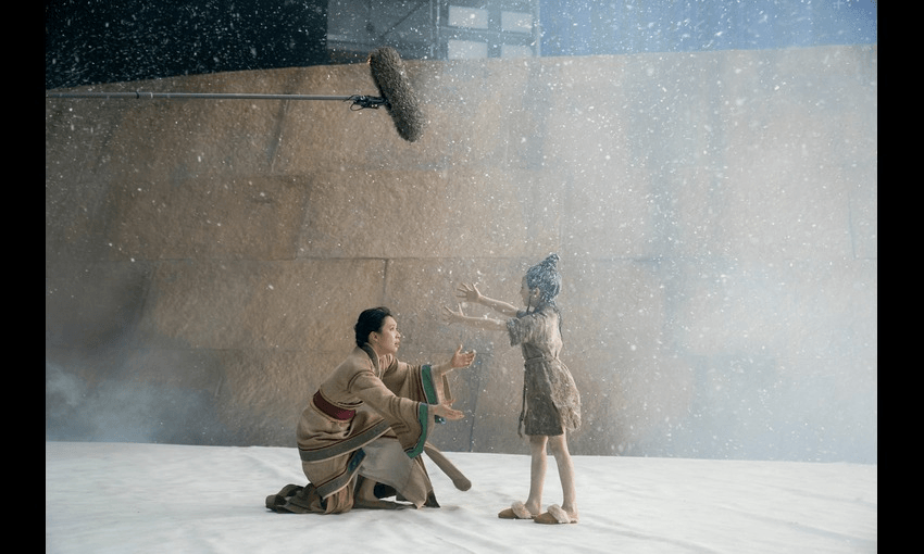 Jess Hong and another actor in historical costumes perform a scene on a snowy set, with one kneeling and the other standing and reaching out; a boom microphone is visible overhead.