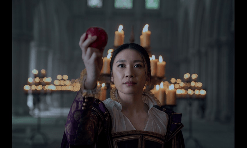 Jess Hong wear a historic costume and holds an apple up on the set of Three Body Problem, there are many candles lit behind her.