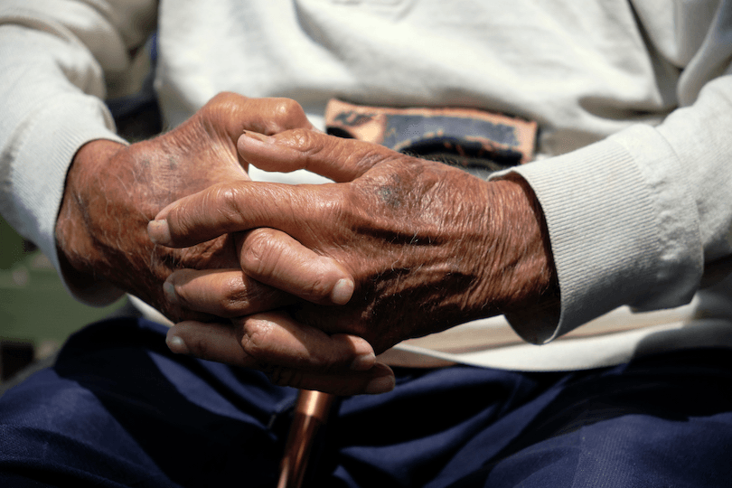 Dark skin toned man with hands clasped. Elderly man sitting with a walking stick with his hands clasped in his lap. Model wears light colored long sleeve jumper and dark blue trousers.