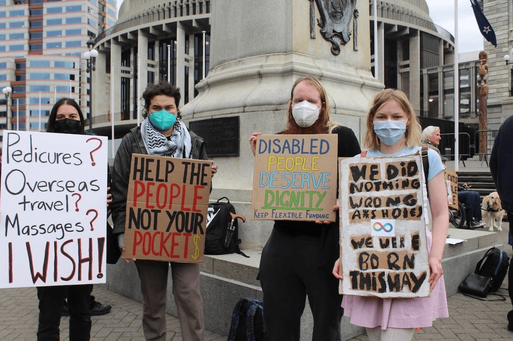 four people wearing masks in front of a statue with signs saying 'pedicures? overseas travel? I WISH) and "Disabled people deserve dignity" and "We did nothing wrong a*seholes, we were born this way"