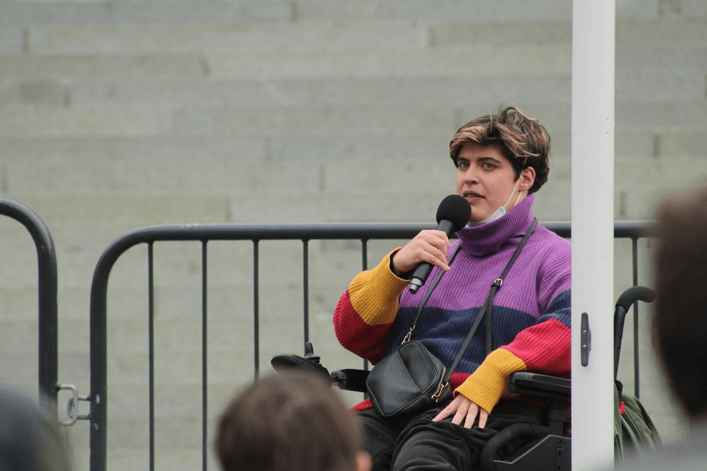 Etta, a person in a wheelchair with short hair wearing a funky purple, red and mustard jumper, is speaking on the parliament forecourt into a microphone