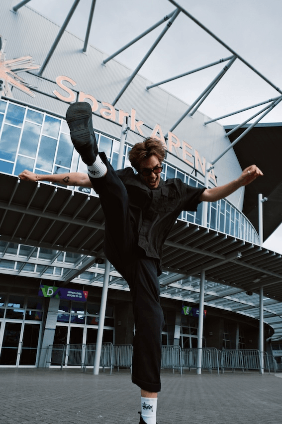 A young man with light brown hair, wearing sunglasses and a black outfit, is performing a high kick in front of Spark Arena. He is mid-air, with one leg raised high and his arms outstretched for balance. The large, modern facade of the arena, with its name visible in the background, creates a dynamic urban setting