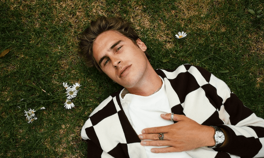 Taylor Roche with light brown hair is lying on grass, wearing a white and brown checkered cardigan over a white shirt. He has his left hand placed on his chest, showcasing a ring and a watch. There are a few white flowers scattered around him on the grass.