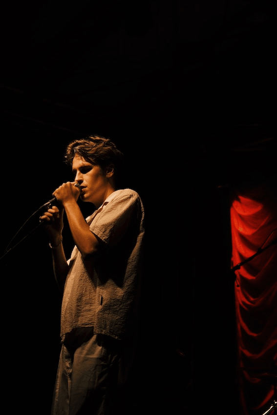 Taylor Roche sings into a microphone on a shadowy stage. He's wearing a white tee and a red stage curtain is seen behind him.