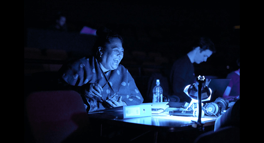 "Anapela Polataivao is seated at a table in a dark room, illuminated by a blue light. She is smiling and appears to be engaged in an activity, with various objects including a water bottle, a small desk lamp, and other items on the table. Another person is visible in the background, working on a laptop."