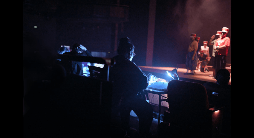 Anapela sits at a desk in the theatre, a blue light shines down on her notes as she reads them, several actors are on stage in front of her.