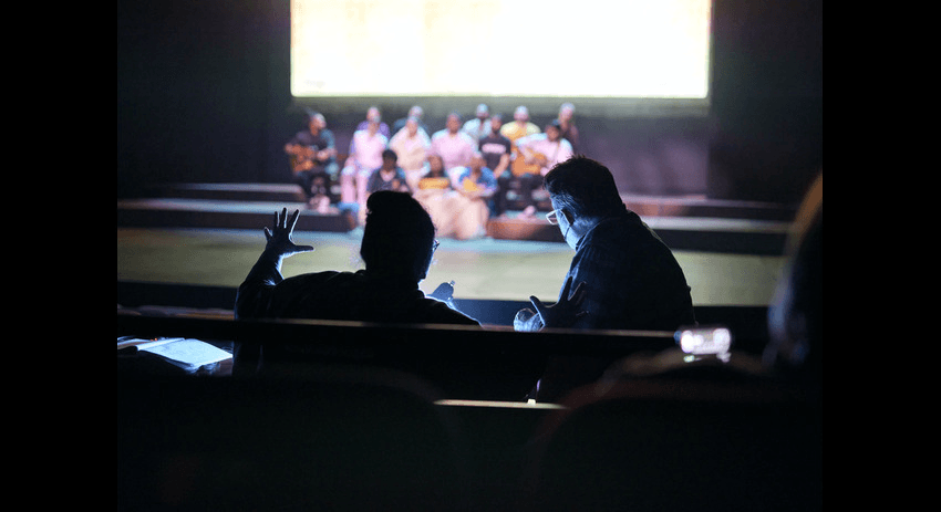 Anapela Polataivao is sitting in a darkened theater, facing the stage with another person. Both are engaged in a conversation, with Anapela gesturing with her hand. On the stage in the background, a group of performers in colorful costumes are seated, illuminated by bright lights.