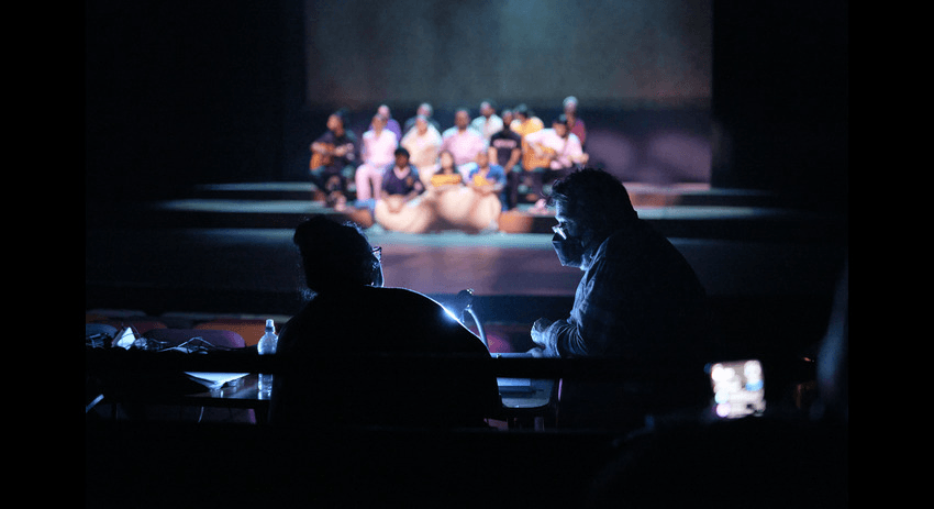 "Anapela Polataivao is sitting in a darkened theater, facing the stage with another person. Both are engaged in a conversation, with Anapela gesturing with her hand. On the stage in the background, a group of performers in colorful costumes are seated, illuminated by bright lights."