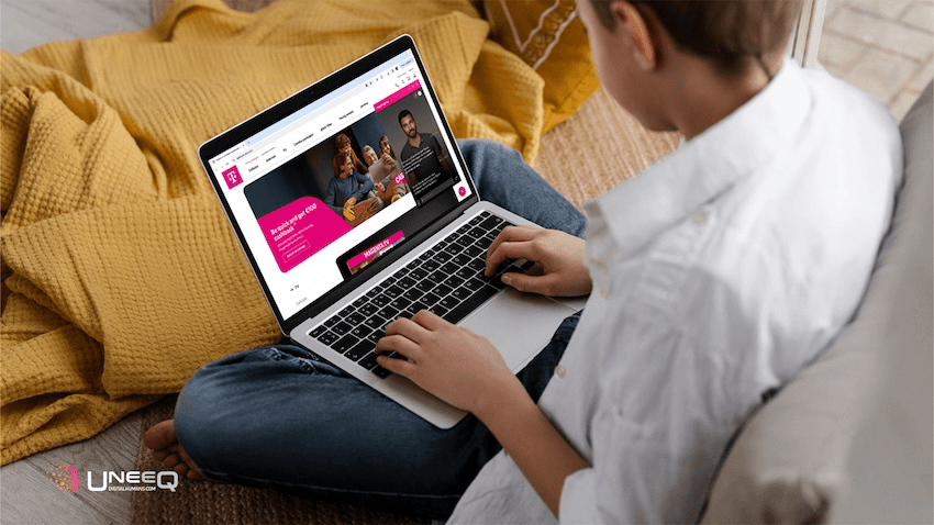 A young person sitting on a cozy, yellow blanket while using a laptop. The laptop screen displays the Deutsche Telekom website featuring a digital assistant named Max, created by UneeQ. The UneeQ logo is visible in the bottom left corner. The setting is a comfortable, well-lit home environment.